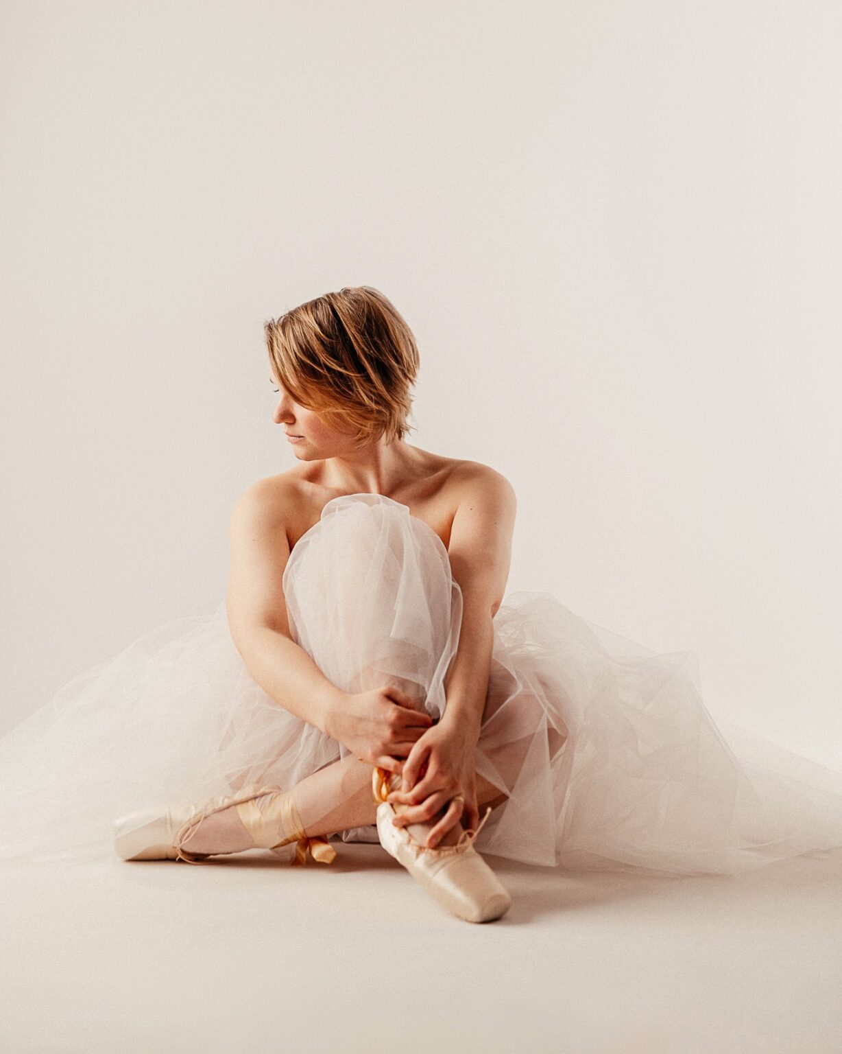 Bild einer Tänzerin in Tüllkleid und Ballettschuhen, die auf dem Boden sitzt und sich zärtlich an ih.